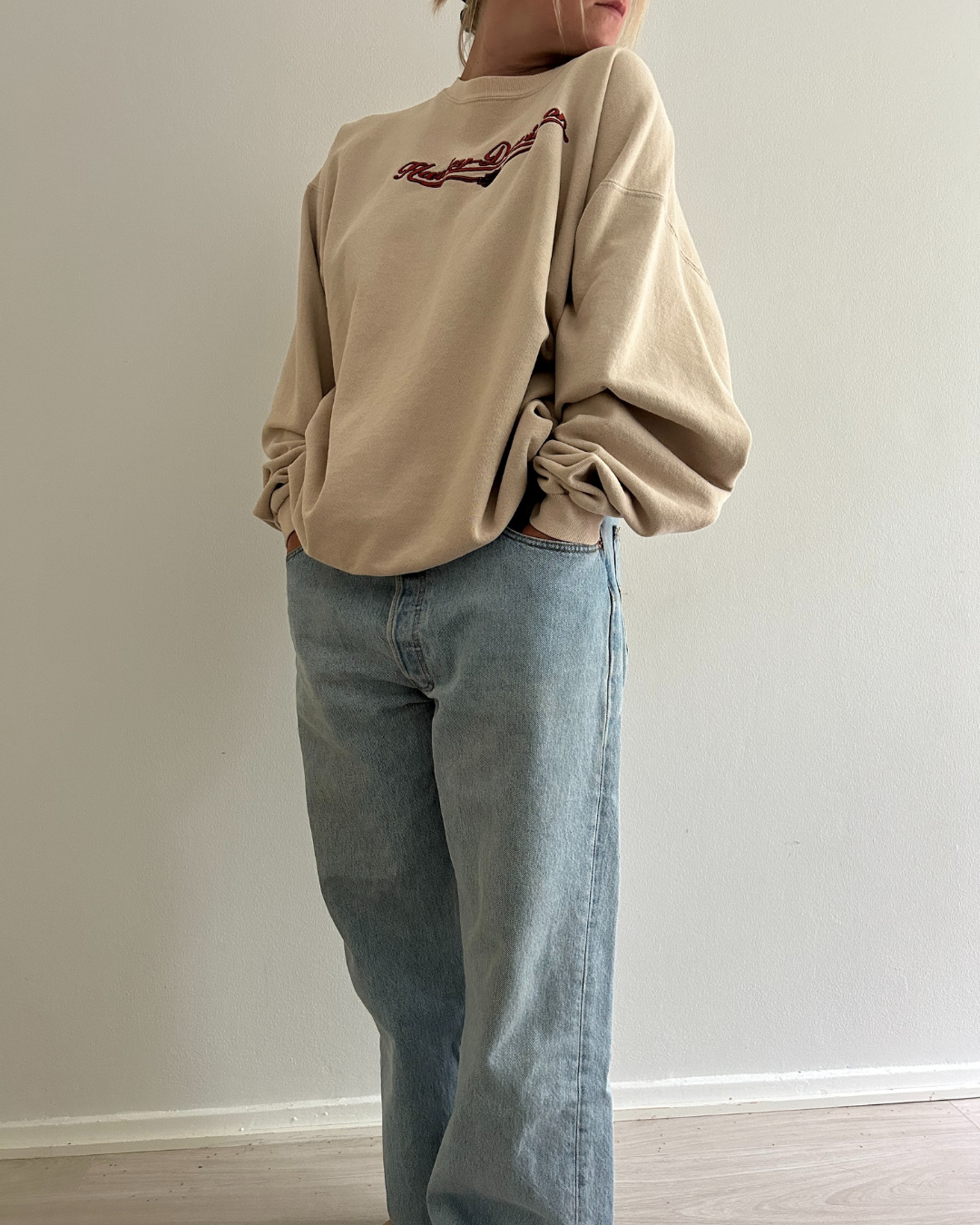 Person wearing a beige sweater with a Harley-Davidson logo and blue jeans against a plain wall.